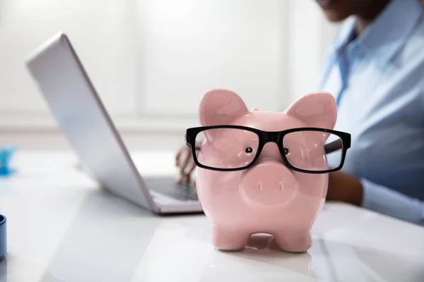 Close-up Of Pink Piggy Bank In Front Of A Business Person Using Laptop At Workplace