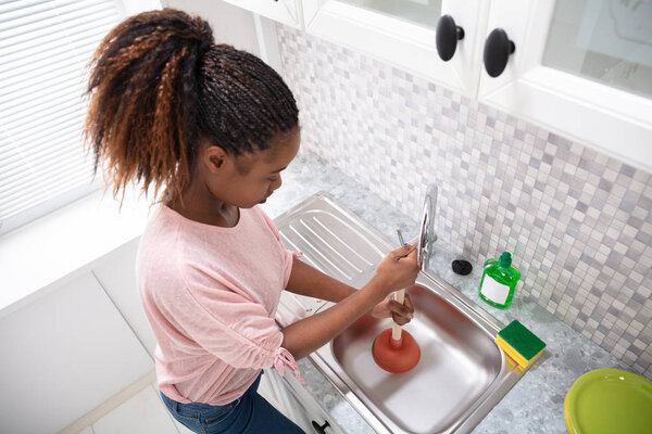 Side View Of A Female Plumber Using Plunger In Kitchen Sink