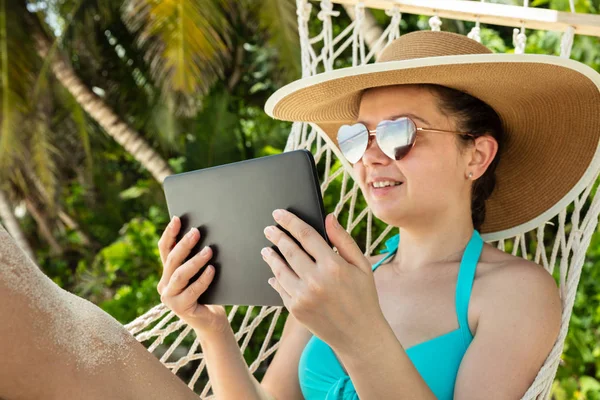 微笑的年轻女子与太阳镜和帽子使用数字平板电脑躺在吊床上在海滩 — 图库照片