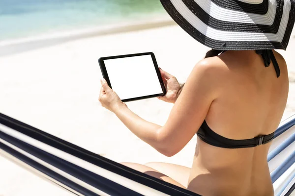在海滩使用数字平板电脑坐在吊床上的妇女坐在吊床上的后视图 — 图库照片