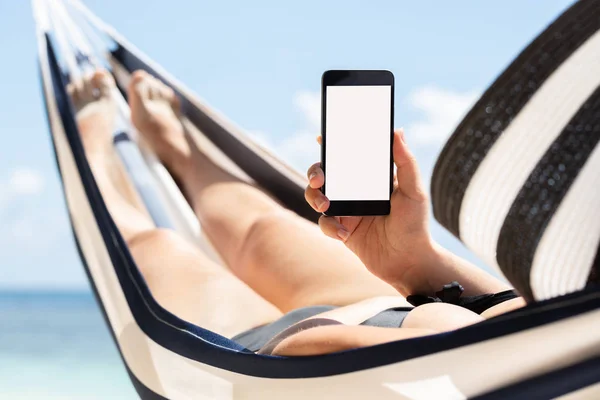 Young Woman Lying On Hammock Using Mobile Phone With White Screen Display Against Blue Sky