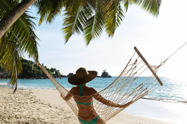 Rear View Of Young Woman In Bikini And Hat Sitting On Hammock On The Beach