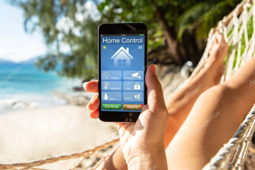 Woman Lying On Hammock Using Smart Home Control On Mobile Phone At Beach