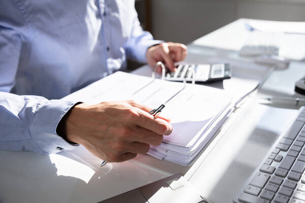 Businessperson Calculating Invoice Using Calculator At Desk