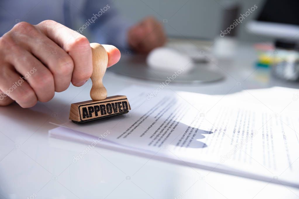 Close-up Of A Person's Hand Stamping With Approved Stamp On Document At Desk