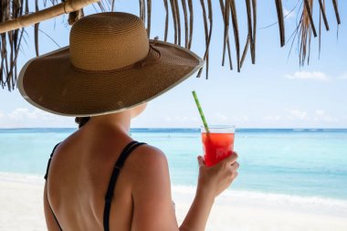Şapkalı, elinde meyve suyu bardağıyla denize bakan bir kadının arka görüntüsü.