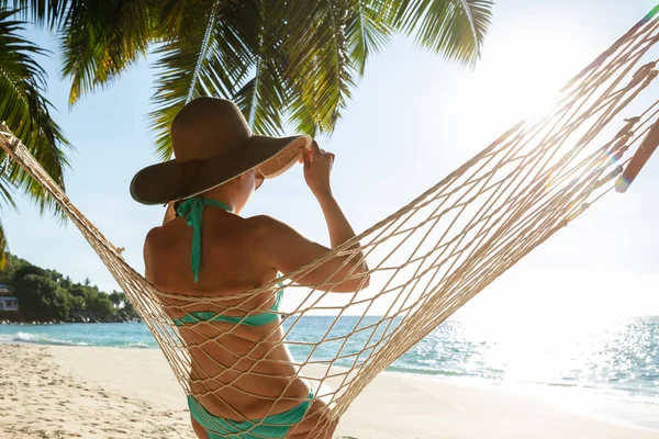 年轻女子在比基尼和帽子坐在海滩上的吊床上的后视图 — 图库照片