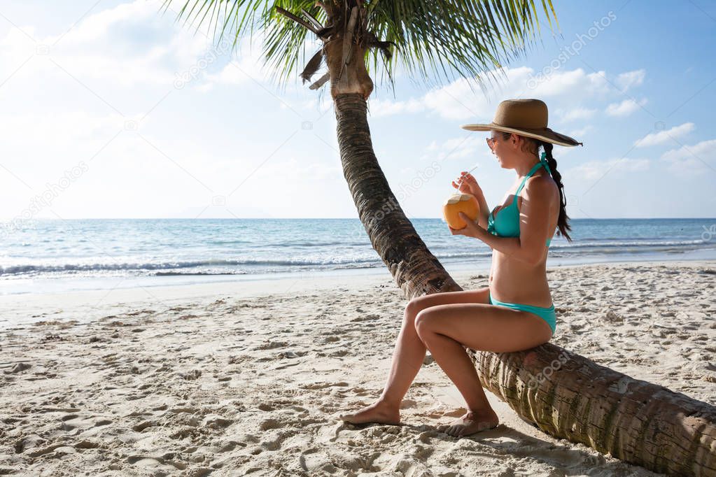 Young Woman In Bikini Sitting On Palm Tree Trunk Drinking The Coconut Water At Beach
