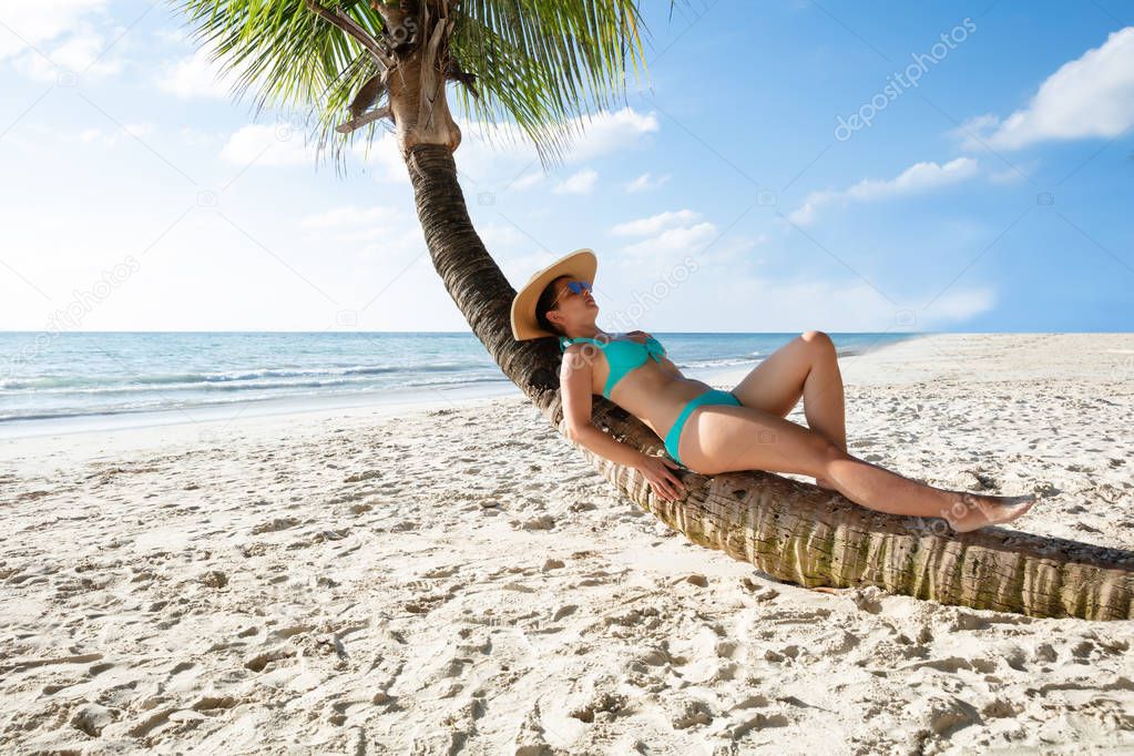 Young Woman In Bikini Relaxing On Crooked Palm Tree At Sandy Beach