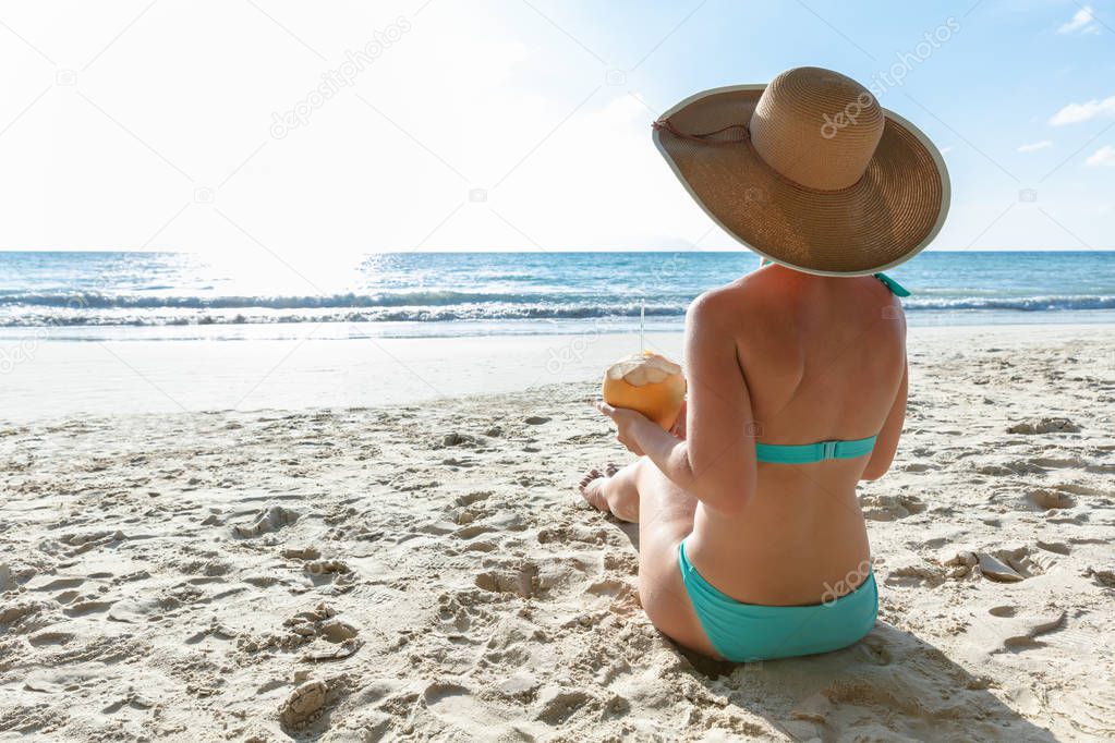 Rear View Of A Woman In Bikini Hat Sitting On Sand Holding Coconut At Beach