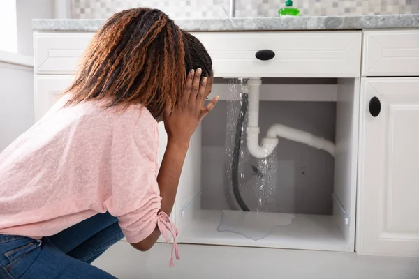 Worried Woman Looking At Leaking Sink Pipe In Kitchen