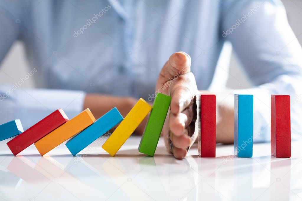 Close-up Of A Businesswoman Hand Stopping Colorful Dominoes From Falling On Office Desk