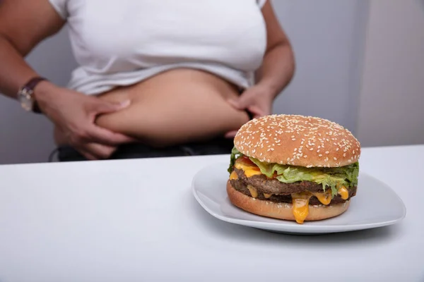 女性の前でおいしいハンバーガー彼女の胃の皮膚をつまむ — ストック写真