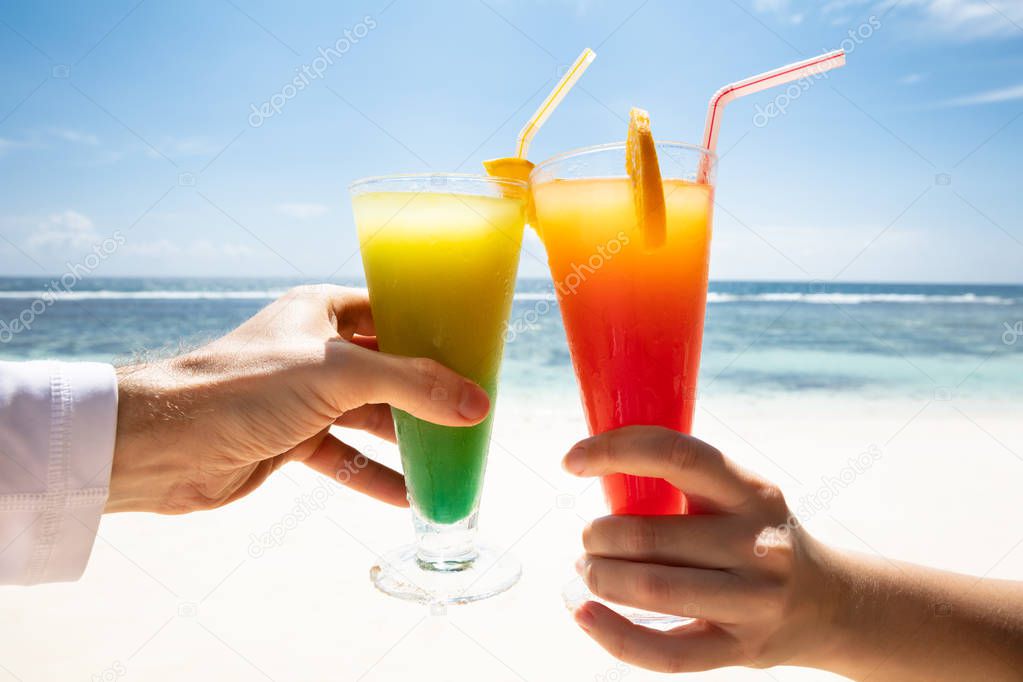 Close-up Of Couple's Hand Toasting The Colorful Cocktail Glasses In Front Of Sea At Beach