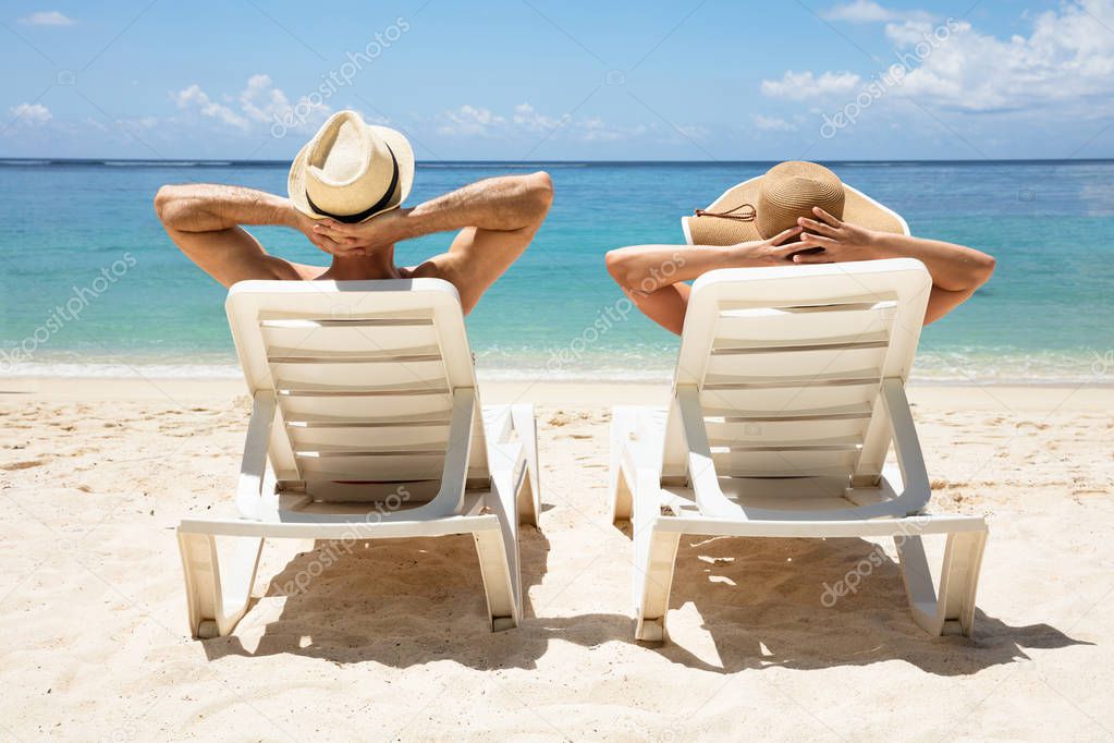 Rear View Of A Young Couple Wearing Hat Relaxing On Deck Chair On Beach