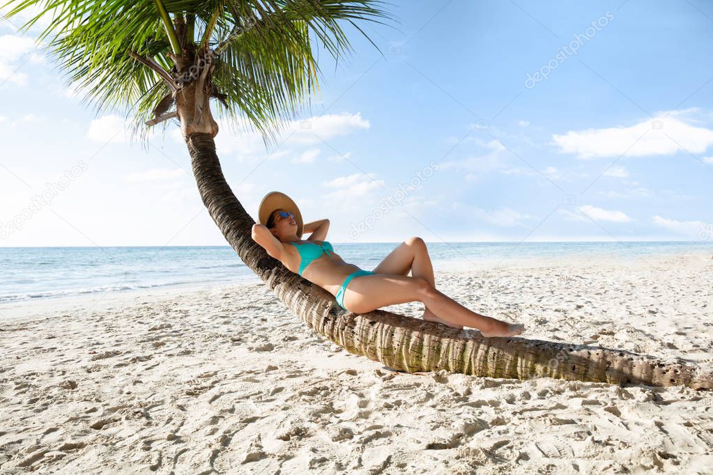 Young Woman In Bikini Relaxing On Crooked Palm Tree At Sandy Beach