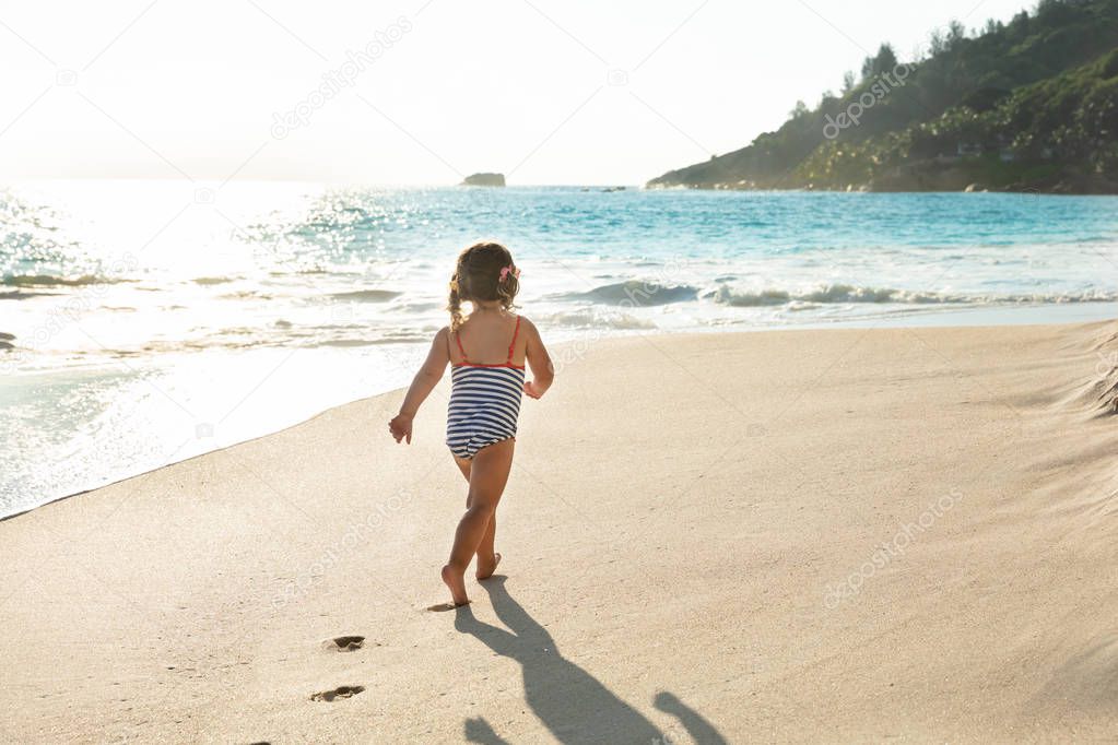 Rear View Of A Toddler Girl In Swimsuit Walking In Sand At Beach