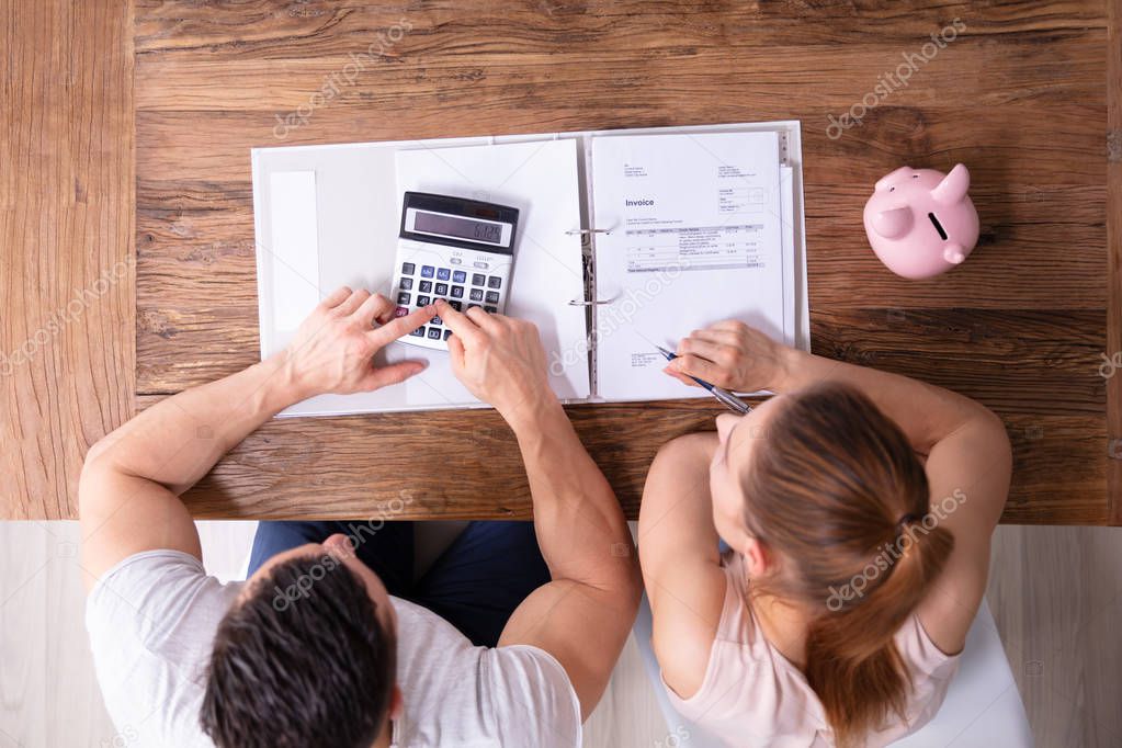 An Overhead View Of Couple Calculating Tax Invoice Using Calculator On Wooden Desk At Home