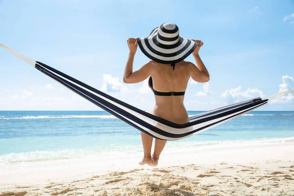 一个穿着比基尼的年轻女子的侧视图 帽子坐在海滩的吊床上 — 图库照片