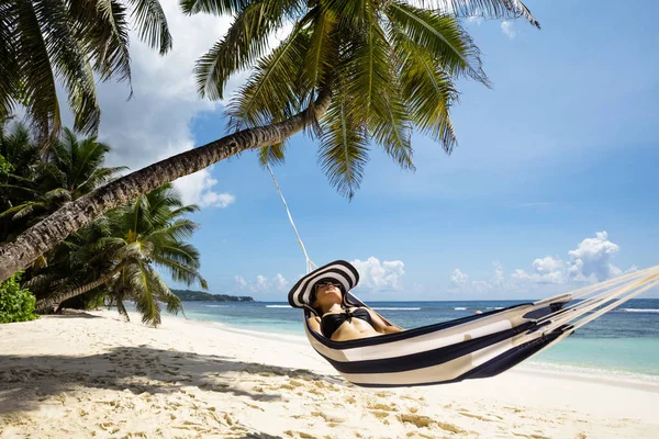 在海岸附近的沙滩上的吊床上 带着条纹帽子放松的年轻女子 — 图库照片