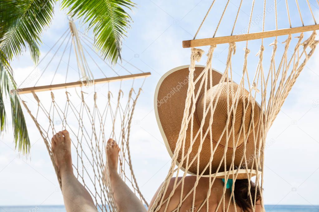 Rear View Of A Young Woman Wearing Hat Relaxing On Hammock Against Blue Sky