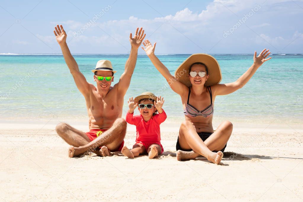 Happy Family Raising Their Hands Sitting On Sand Enjoying The Vacation At Beach