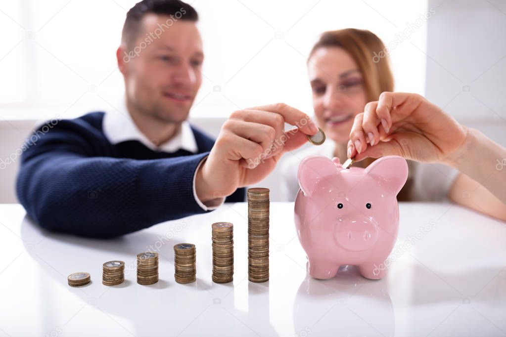 Couple Inserting Coin In Piggybank Near Increasing Profit Over Desk