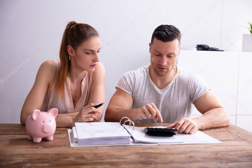 Couple Calculating Bills Using Calculator Near Piggy Bank At Workplace