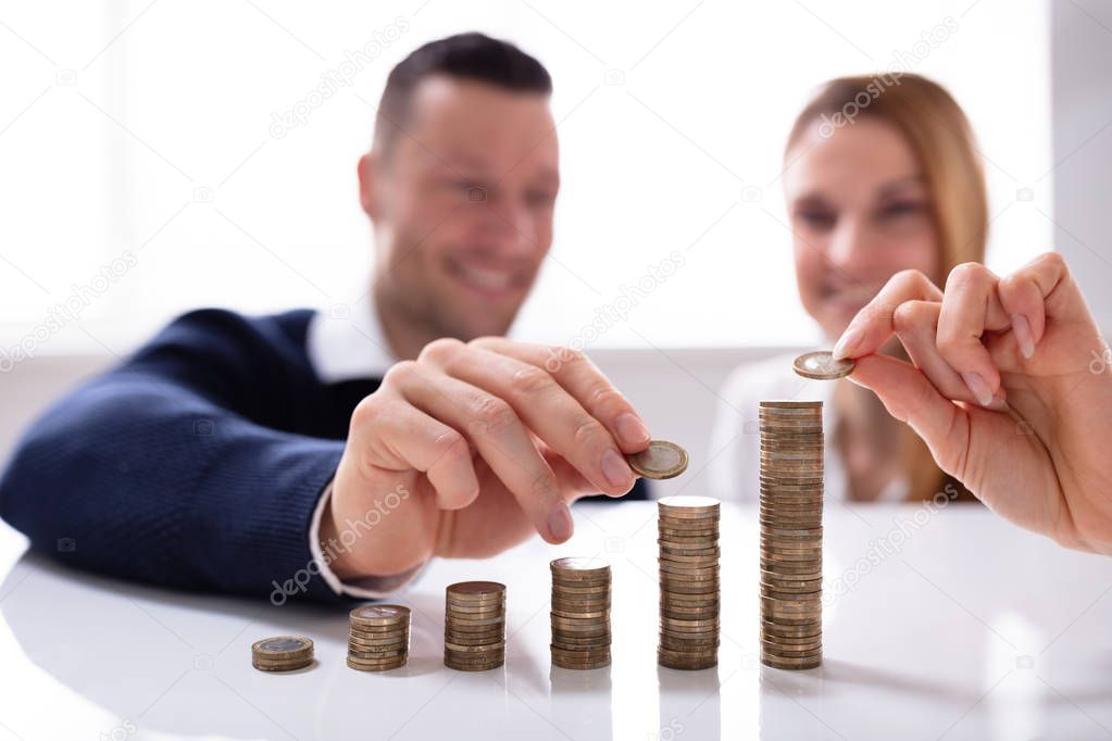Couple's Hand Stacking Coins Over White Desk In Office