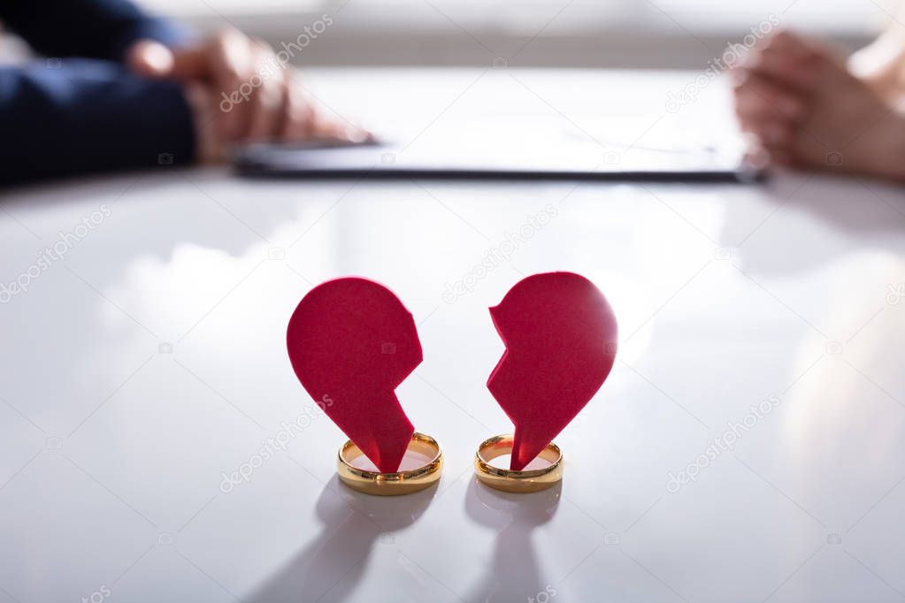 Close-up Of Broken Red Heart And Weeding Rings Over White Desk