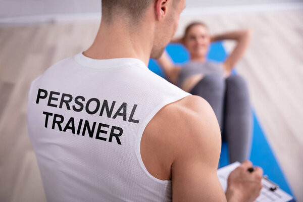 Personal Trainer Looking At Young Woman Doing Exercise On Yoga Mat