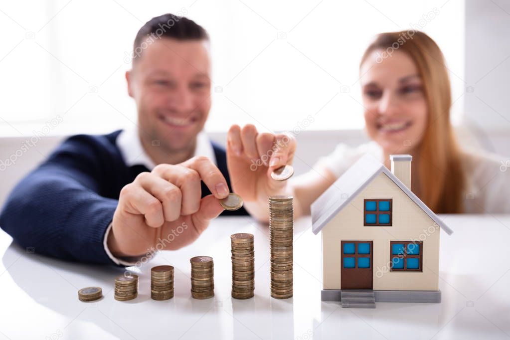 Couple Placing The Coin On Top Of The Increasing Stack Of Coins Near House Model