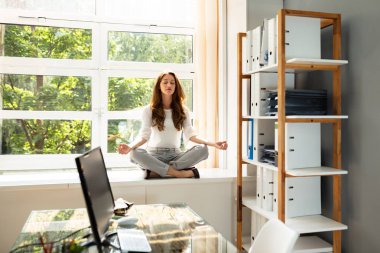 Relaxed Young Businesswoman Meditating In Lotus Position Over Cabinet Near Window clipart