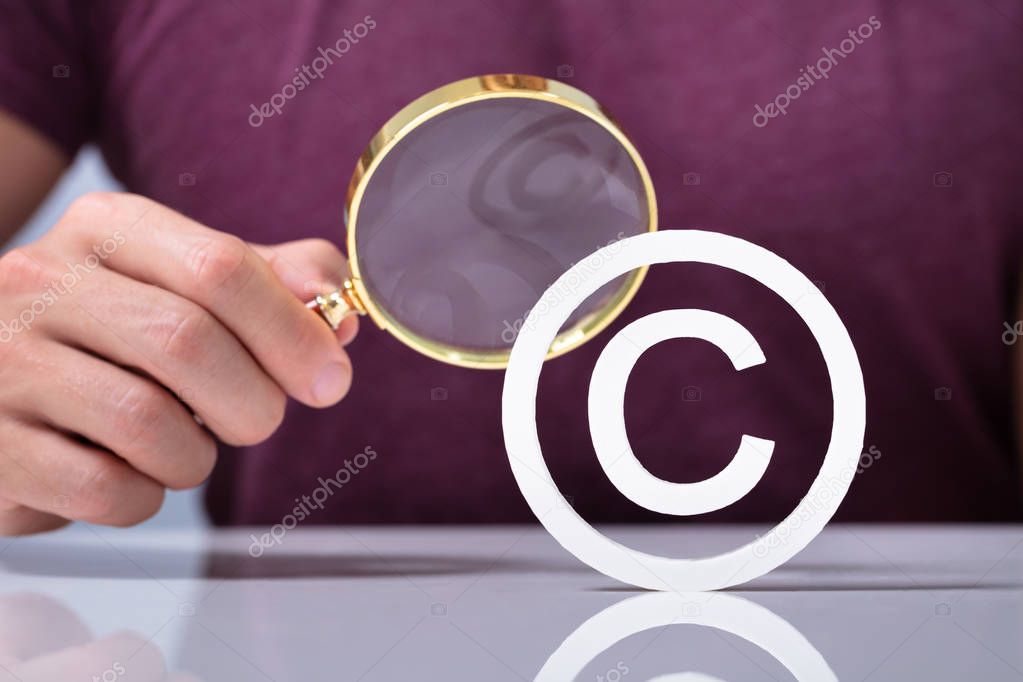 A Person's Hand Holding Magnifying Glass Looking Copyright Symbol Icon Over Desk