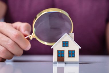 Human Hand Holding Magnifying Glass Looking House Model Over White Desk clipart