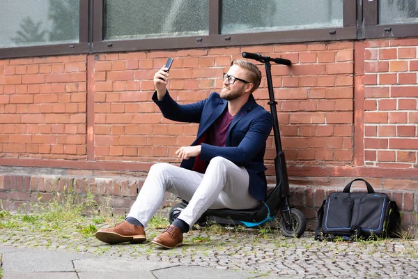 Uśmiechnięty Biznesmen Siedzi Skuter Elektryczny Pobliżu Brick Wall Biorąc Selfie — Zdjęcie stockowe