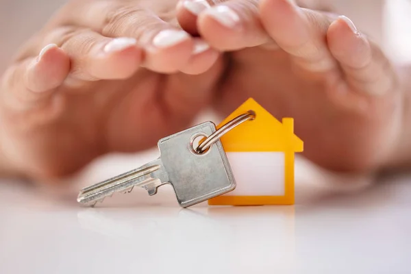 Close-up Of Person\'s Hand Protecting House Key With Keychain On White Desk