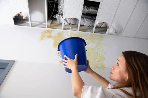 担心的女人拿着一个蓝色的桶在走廊的泄漏天花板下 — 图库照片