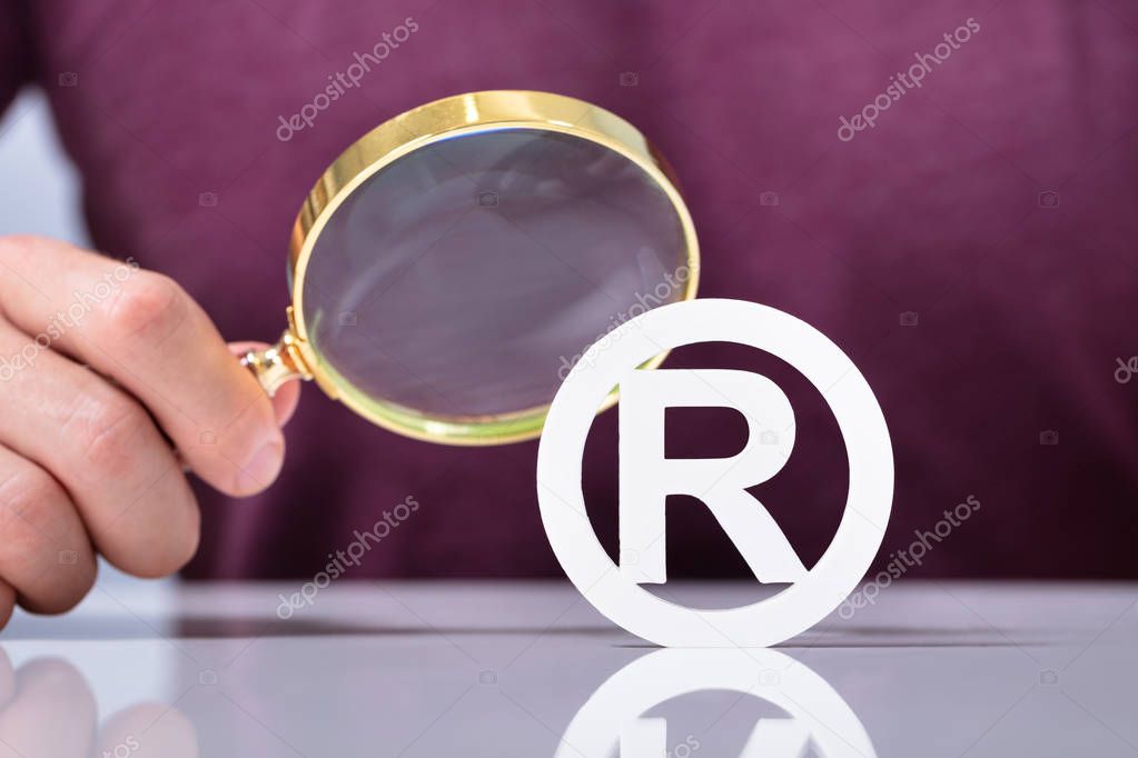 Human Hand's Holding Magnifier Looking Registered Trademark Icon Over Reflective Desk