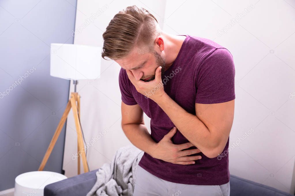 Young Man Suffering From Nausea Standing In Front Of Sofa At Home