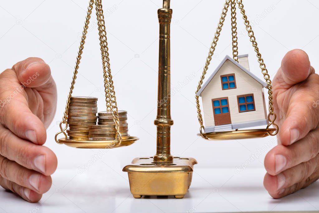 Close-up Of A Person's Hand Protecting Justice Scale With Stacked Coins And House Model