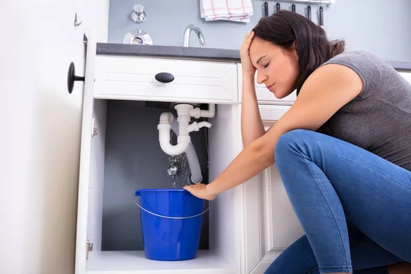 青いバケツのシンクから漏れる水を集める心配した女性 — ストック写真