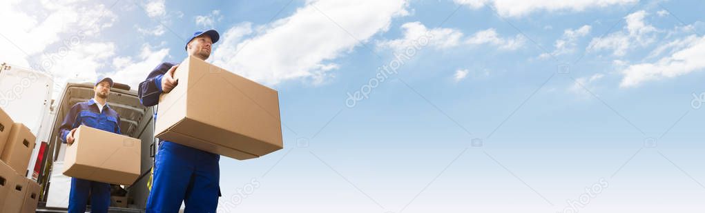 Low Angle View Of Two Young Delivery Man Carrying Cardboard Box In Front Of Truck