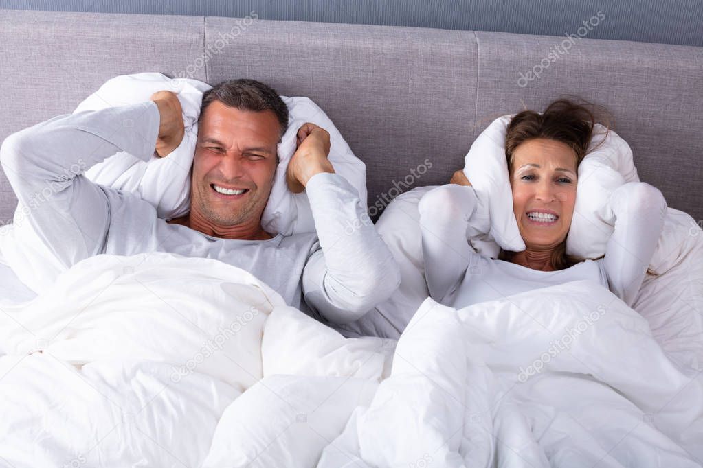 Mature Couple Disturbed By Noise Covering Their Ears With Pillow On Bed