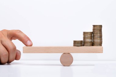 Close-up Of A Businessperson's Hand Balancing Stacked Coins On Wooden Seesaw With Finger Over Desk clipart