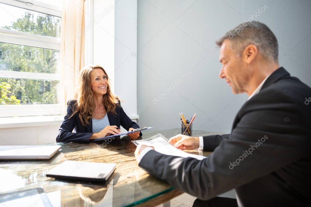 Mature Businesswoman Taking An Interview Of Man Over The Wooden Desk In Office