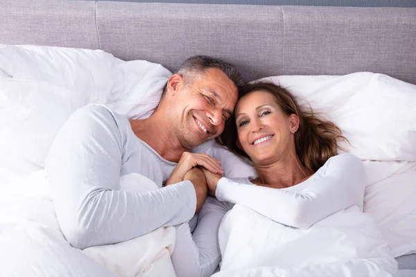 自宅でベッドに横たわりながら お互いに手をつないで微笑む成熟したカップル — ストック写真
