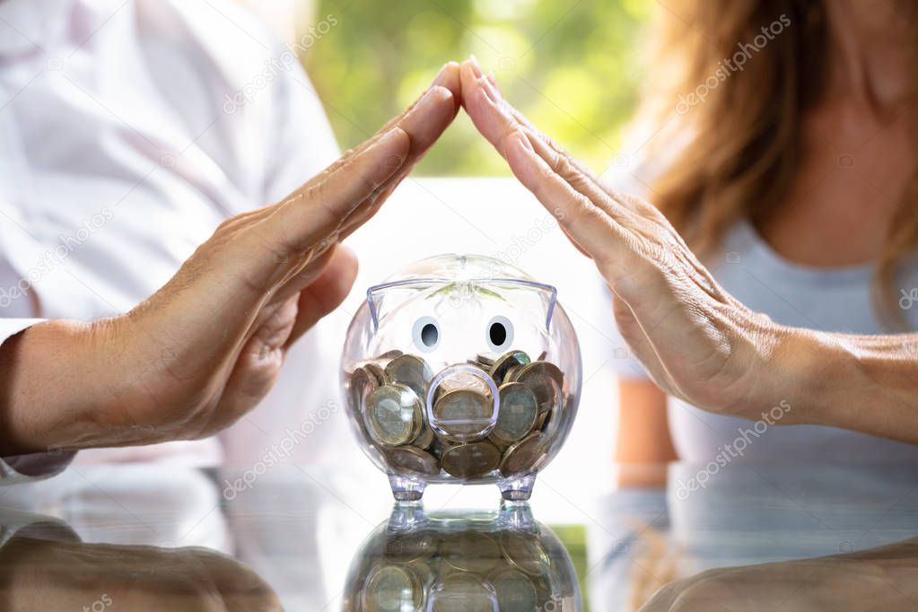 Close-up Of A Human Hand Protecting Transparent Piggy Bank On Reflective Desk