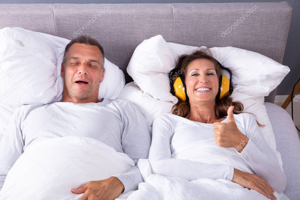 Happy Mature Woman Covering Her Ears With Headphone Showing Thumb Up Sign While Man Snoring