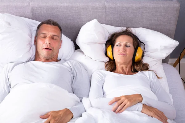 男がベッドでいびきをかいている間 ヘッドフォンで彼女の耳を覆う成熟した女性 — ストック写真